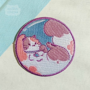 May include: An embroidered patch featuring a white and pink cat inside a bubble with a blue background. The cat is wearing a pink and white hat and has pink ears.