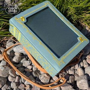 Könnte beinhalten: Eine teal-farbene Handtasche mit einem transparenten Fenster, goldfarbener Blumenstickerei und einem braunen Schulterriemen. Die Tasche hat eine rechteckige Form und liegt auf Steinen und Grün. Der Markenname "calico cocoa" ist sichtbar.