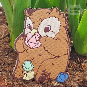 Peut inclure: Hibou brodé marron tenant une pierre précieuse rose dans ses pattes. Le hibou est assis sur un tas de pierres précieuses colorées, dont une pierre précieuse bleue, verte et jaune.