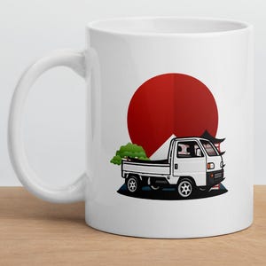 Op de afbeelding: Witte keramische mok met een Japanse illustratie. Het ontwerp toont een rode zon, een witte vrachtwagen en een groene boom. De mok heeft een gebogen handvat en staat op een houten oppervlak.