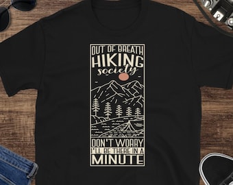 Op de afbeelding: Zwart T-shirt met een grafisch ontwerp van een berglandschap met de tekst "Out of Breath Hiking Society Don't Worry I'll be there in a minute."