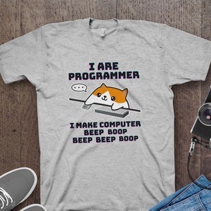 Op de afbeelding: Een grijs T-shirt met de tekst "I ARE PROGRAMMER" en een cartoonkat die op een toetsenbord typt. De tekst onder de kat zegt "I MAKE COMPUTER BEEP BOOP BEEP BEEP BOOP".