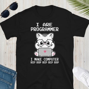 Op de afbeelding: Zwart T-shirt met een witte cartoonkat die een bril draagt en een laptop gebruikt. De tekst "I are programmer" en "I make computer beep boop beep beep boop" staat op het T-shirt gedrukt.