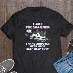 Op de afbeelding: Zwart T-shirt met een witte en zwarte cartoonkat die een roze headset draagt en op een toetsenbord typt. De tekst op het shirt luidt "I are programmer I make computer beep boop beep beep boop".