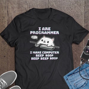 Op de afbeelding: Zwart T-shirt met de tekst "I ARE PROGRAMMER" en een cartoonkat die een toetsenbord bespeelt. Onder de kat staat de tekst "I MAKE COMPUTER BEEP BOOP BEEP BOOP BOOP". Het shirt ligt op een houten oppervlak met schoenen, een spijkerbroek en een camera.