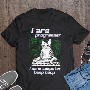 Op de afbeelding: Zwart T-shirt met een witte kat die een groen shirt draagt en op een toetsenbord typt. De tekst op het T-shirt luidt "I are programmer I make computer beep boop".