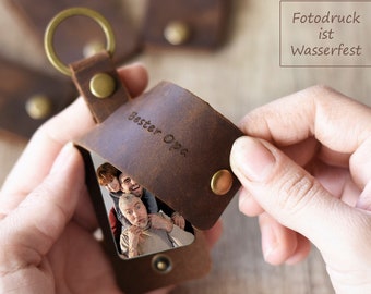 May include: A brown leather keychain with a photo insert. The keychain has a metal ring and a snap closure. The photo insert shows a family of four. The text "Bester Opa" is engraved on the leather. The text "Fotodruck ist Wasserfest" is printed on a white background.