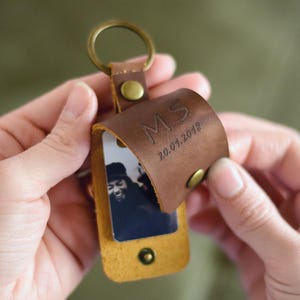 Könnte beinhalten: Ein brauner Leder-Schlüsselanhänger mit Messingring und Druckknopfverschluss. Der Schlüsselanhänger hat eine kleine Klappe mit den Initialen "MS" und dem Datum "20.01.2018" eingraviert. Ein Schwarz-Weiß-Foto ist sichtbar.