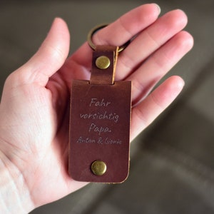 K&ouml;nnte beinhalten: Ein rechteckiger Schl&uuml;sselanh&auml;nger aus dunkelbraunem Leder mit einem messingfarbenen Metallring und zwei messingfarbenen Druckkn&ouml;pfen. Die Lederoberfl&auml;che ist mit dem Text graviert: Fahr vorsichtig Papa. Anton & Liorie.
