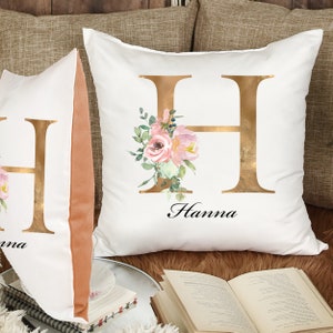 May include: Two white pillow covers with gold floral designs. The pillows feature a large gold letter "H" with pink and white flowers and greenery. The name "Hanna" is written in black script below the letter.