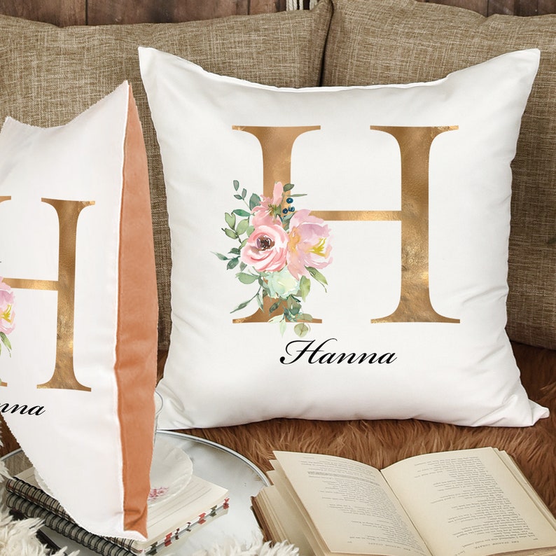 May include: Two white pillow covers with gold floral designs. The pillows feature a large gold letter "H" with pink and white flowers and greenery. The name "Hanna" is written in black script below the letter.