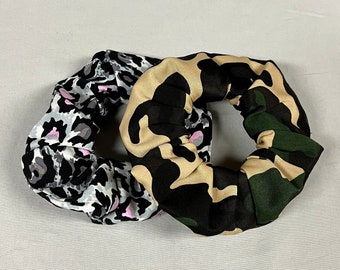 May include: Two fabric hair scrunchies. One is black, green, and beige camouflage print. The other is gray and white leopard print with pink accents.