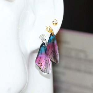 May include: A pair of dangle earrings featuring a pink and blue butterfly wing design. The wings are translucent and have gold flecks. The earrings are attached to silver hooks.