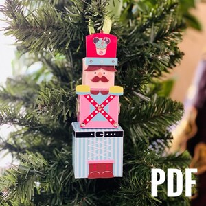 Puede incluir: Un adorno de cascanueces de madera con un diseño rosa, azul y blanco. El cascanueces tiene un sombrero a rayas rojo y blanco con una cabeza de Mickey Mouse y un cupcake en la parte superior. El adorno está colgado de un árbol de Navidad.