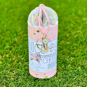 Puede incluir: Un tubo de papel rosa y blanco con un dije dorado y una margarita. El tubo tiene una parte superior plisada y el texto "Nice", "Stylin", "Cheer" y "Dude" en el lateral.