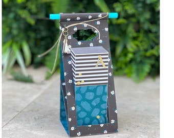 Pode incluir: Uma sacola de presente preta e branca com uma caixa de padrão azul esverdeado e branco dentro. A sacola tem um tassel branco e um padrão floral branco. A caixa tem um padrão listrado preto e branco e a letra "A" em dourado.