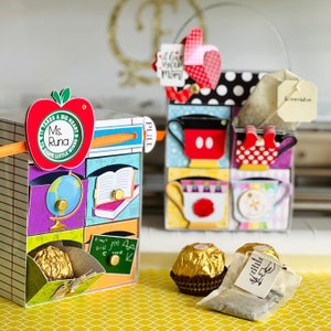 Puede incluir: Una caja de cartón colorida con una lengüeta de extracción, decorada con una manzana roja, un globo terráqueo, un libro y el texto "It Takes A Big Heart To Shape Little Minds Ms. Rund". La caja está llena de pequeños regalos, incluyendo un caramelo de chocolate. También se puede ver una bolsita de té en la imagen.