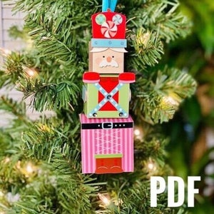 Puede incluir: Un adorno de cascanueces de papel artesanal colorido con un sombrero rojo, una chaqueta verde y pantalones rosas. El adorno está colgado de un árbol de Navidad.