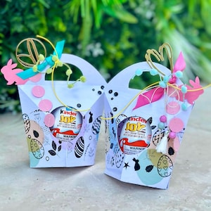 Puede incluir: Dos cajas de regalo blancas con temática de Pascua con asas, cada una con un huevo Kinder Joy. Las cajas están decoradas con colores pastel, diseños de huevos de Pascua y recortes de conejitos rosas. Adornos dorados y rosas adornan las asas.