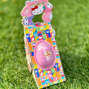 Puede incluir: Una colorida caja de regalo con un huevo Play-Doh rosa y una parte superior decorativa con personajes de dibujos animados y el nombre "Sammy". La caja tiene un patrón vibrante de personajes de dibujos animados y formas geométricas. La caja está sobre hierba verde.