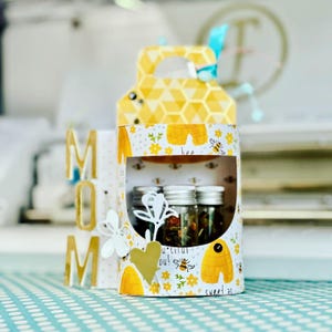 Puede incluir: Una caja de regalo decorativa con un patrón de panal de abeja amarillo y un asa. La caja presenta una ventana que revela pequeños frascos de vidrio. La caja está decorada con ilustraciones de abejas y flores, y la palabra "MOM" se muestra al lado.