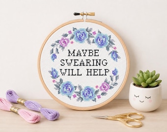 May include: A circular wooden embroidery hoop containing white fabric with a cross-stitch design. The design features a floral wreath of blue and purple roses surrounding the black text that reads, MAYBE SWEARING WILL HELP, in a block font.