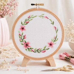 Könnte beinhalten: Ein runder Stickrahmen aus Holz mit weißem Aida-Stoff, der ein Kreuzstichmuster eines Blumenkranzes zeigt. Der Kranz besteht aus rosa Blüten und grünen Blättern, die kreisförmig angeordnet sind. Der Rahmen steht auf einem kleinen Holzständer.