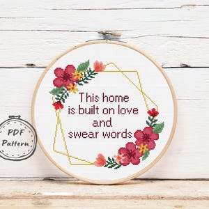 Puede incluir: Patrón de punto de cruz con una corona de flores rojas y amarillas y formas geométricas doradas. El texto dice "Esta casa está construida sobre el amor y las palabrotas".