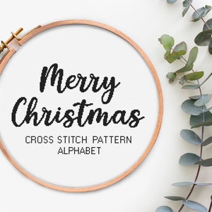 Op de afbeelding: Een houten borduurring met witte Aida-stof erin. De stof heeft een zwart kruissteekpatroon met de tekst "Merry Christmas" en "Cross Stitch Pattern Alphabet".