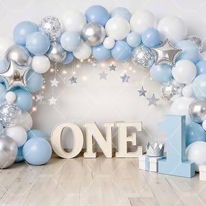 May include: A celebratory first birthday backdrop with a balloon arch in shades of blue, white, and silver. The word "ONE" is displayed in large letters, with a number "1" and gift boxes. Silver stars and fairy lights enhance the festive theme.