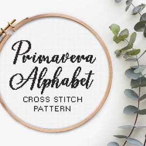 May include: A cross-stitch pattern in a wooden embroidery hoop. The text "Primavera Alphabet" is stitched in black thread on white fabric. The words "Cross Stitch Pattern" are below. Green leaves are in the background.