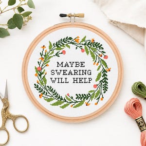 Könnte beinhalten: Ein runder Stickrahmen aus Holz mit weißem Stoff, der ein Kreuzstichmotiv aus einem grünen Blattkranz mit kleinen orangefarbenen und rosafarbenen Blüten zeigt. Der Text in der Mitte lautet MAYBE SWEARING WILL HELP in schwarzen Buchstaben.