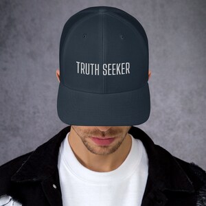 May include: A navy blue mesh trucker hat with a white embroidered design that says "TRUTH SEEKER".