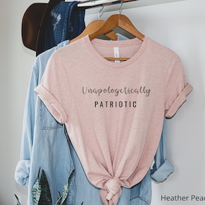 May include: A heather peach colored t-shirt with the text "Unapologetically PATRIOTIC" printed in black.