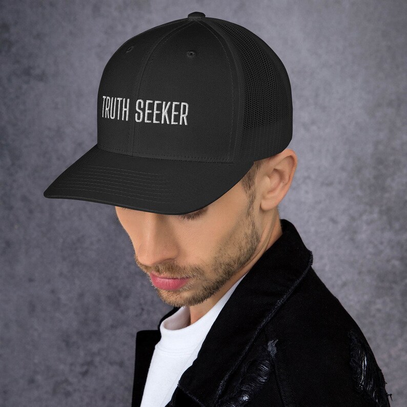 May include: A black trucker hat with a black mesh back. The hat has white embroidered text that reads "TRUTH SEEKER".
