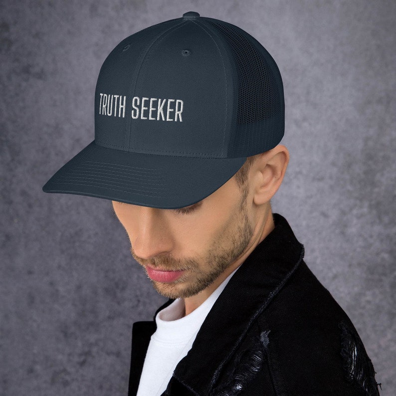May include: A navy blue trucker hat with a black mesh back. The hat has white embroidered text that reads "TRUTH SEEKER".