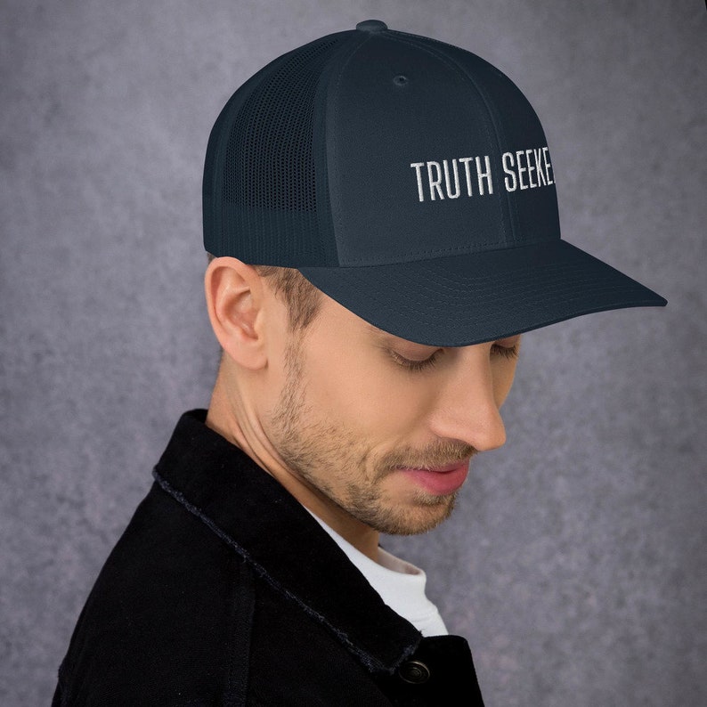 May include: A navy blue trucker hat with a black mesh back. The hat has white embroidered text that reads "TRUTH SEEKER".