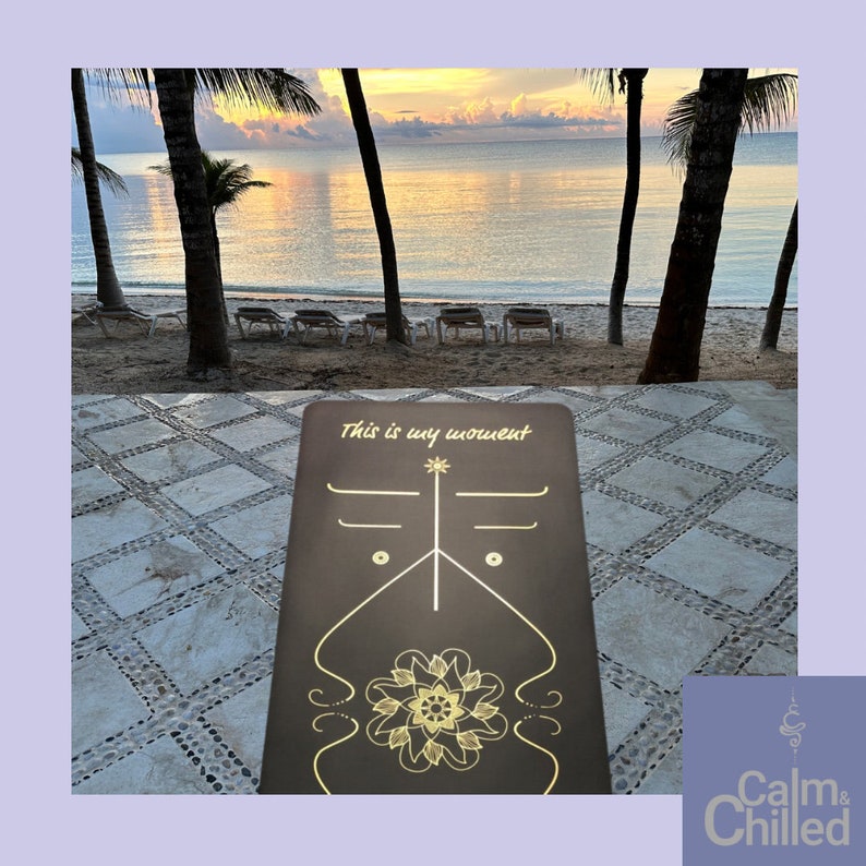 May include: A brown yoga mat with gold lettering that says "This is my moment" and a gold mandala design. The mat is on a tiled surface in front of a beach scene with palm trees and a calm ocean.