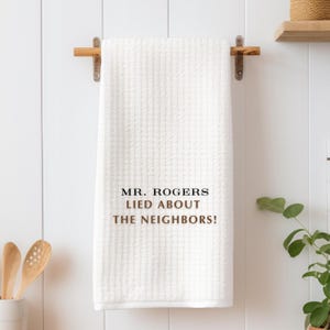 Pu&ograve; includere: Asciugamano da cucina bianco con il testo "MR. ROGERS LIED ABOUT THE NEIGHBORS!" in lettere nere e marroni.