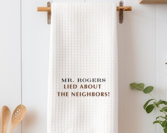 May include: White kitchen towel with the text "MR. ROGERS LIED ABOUT THE NEIGHBORS!" in black and brown lettering.
