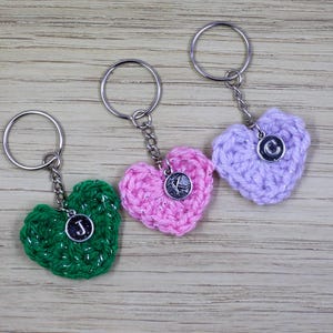 May include: Three handmade heart-shaped keychains in green, pink, and lavender. Each keychain features a crocheted heart with a silver-tone initial charm and a metal keyring. The keychains are displayed on a light wood surface.