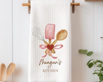 May include: White kitchen towel with a pink spatula, wooden spoon, and whisk tied together with a pink ribbon and a red heart. The text "Megan's Kitchen" is printed on the towel.