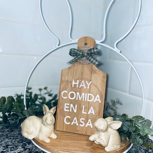 May include: White metal bunny-shaped stand with a wooden cutting board that says "Hay comida en la casa" and two small white bunny figurines on a wooden base.