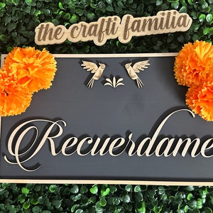 May include: A black wooden sign with the text "Recuerdame" in a decorative font. The sign is decorated with two small wooden birds and orange marigold flowers. The sign also has a smaller wooden sign above it that says "the crafti familia".