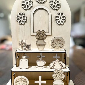 May include: A wooden Day of the Dead altar with multiple shelves. It features laser-cut decorations including flowers, skulls, crosses, a candle, a cup of coffee, and a vase of flowers. The altar is made of light-colored wood.