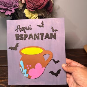 May include: A square purple wooden sign featuring the text "Aqui ESPANTAN" in black lettering, decorated with four black bat silhouettes and a central brown clay-style mug illustration with yellow, pink, and light blue accents.