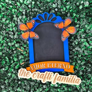 May include: A blue wooden frame with a chalkboard insert and two orange butterflies. The frame has a scalloped top and a banner that reads "AMOR ETERNO". Below the frame is a wooden sign that reads "the crafty familia".