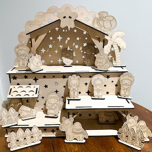 May include: A wooden nativity scene with laser-cut figures and a stable. The stable has a star-shaped cutout and is decorated with star cutouts. The figures include Mary, Joseph, baby Jesus, and various animals. The scene is set on a wooden table.