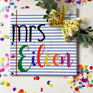 May include: A personalized notebook-style tile with the name "Eileen" in rainbow colors and the title "Mrs." in black. The tile has blue lines and a red margin. A yellow ribbon with school-themed patterns and greenery adorns the top right corner.