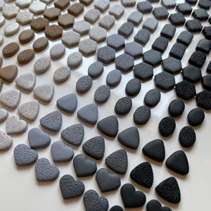 May include: A collection of small, textured ceramic shapes in various shades of gray, from light to dark. The shapes include circles, ovals, hexagons, triangles, and hearts.